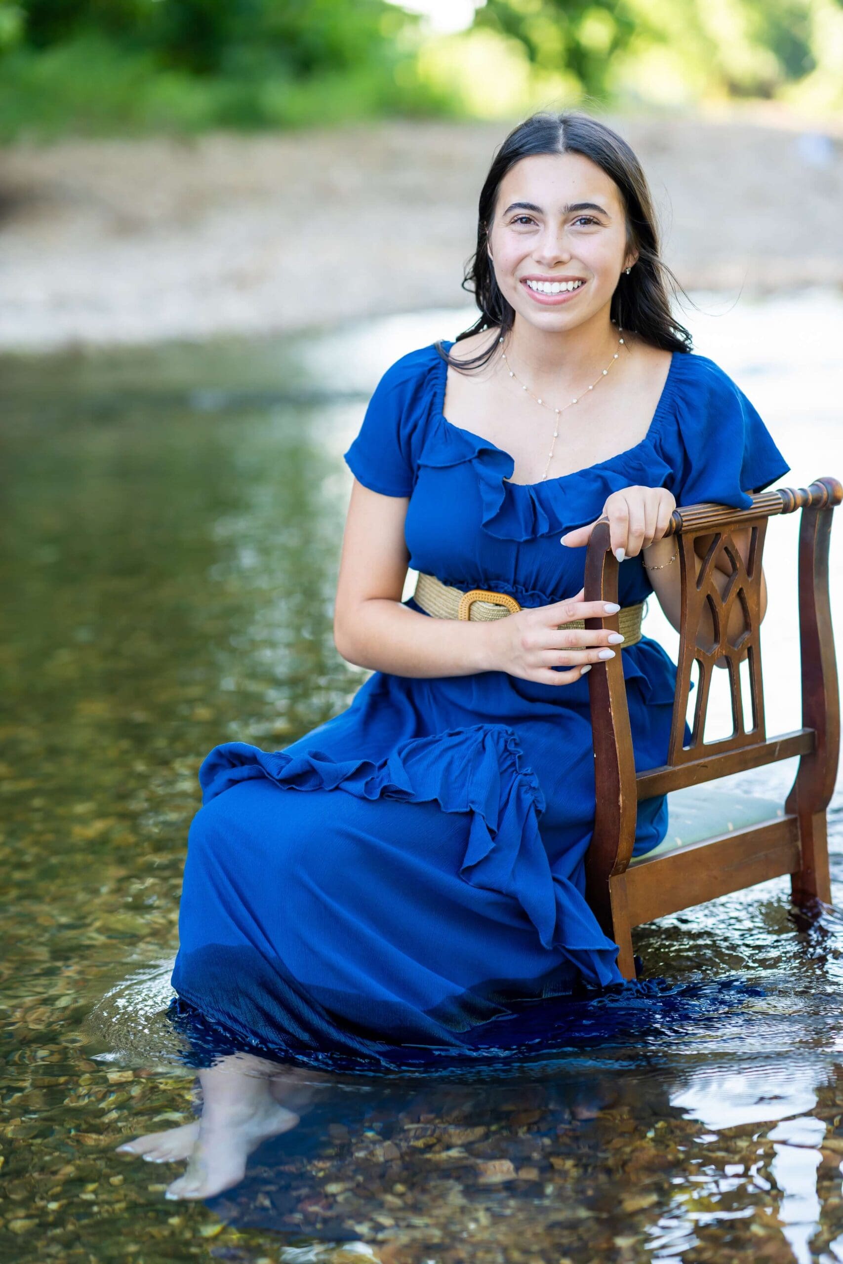 Example Shoot Title Tag An image captured by an Arkansas senior photographer of a high school senior in a blue dress sitting on a chair in a shallow river smiling