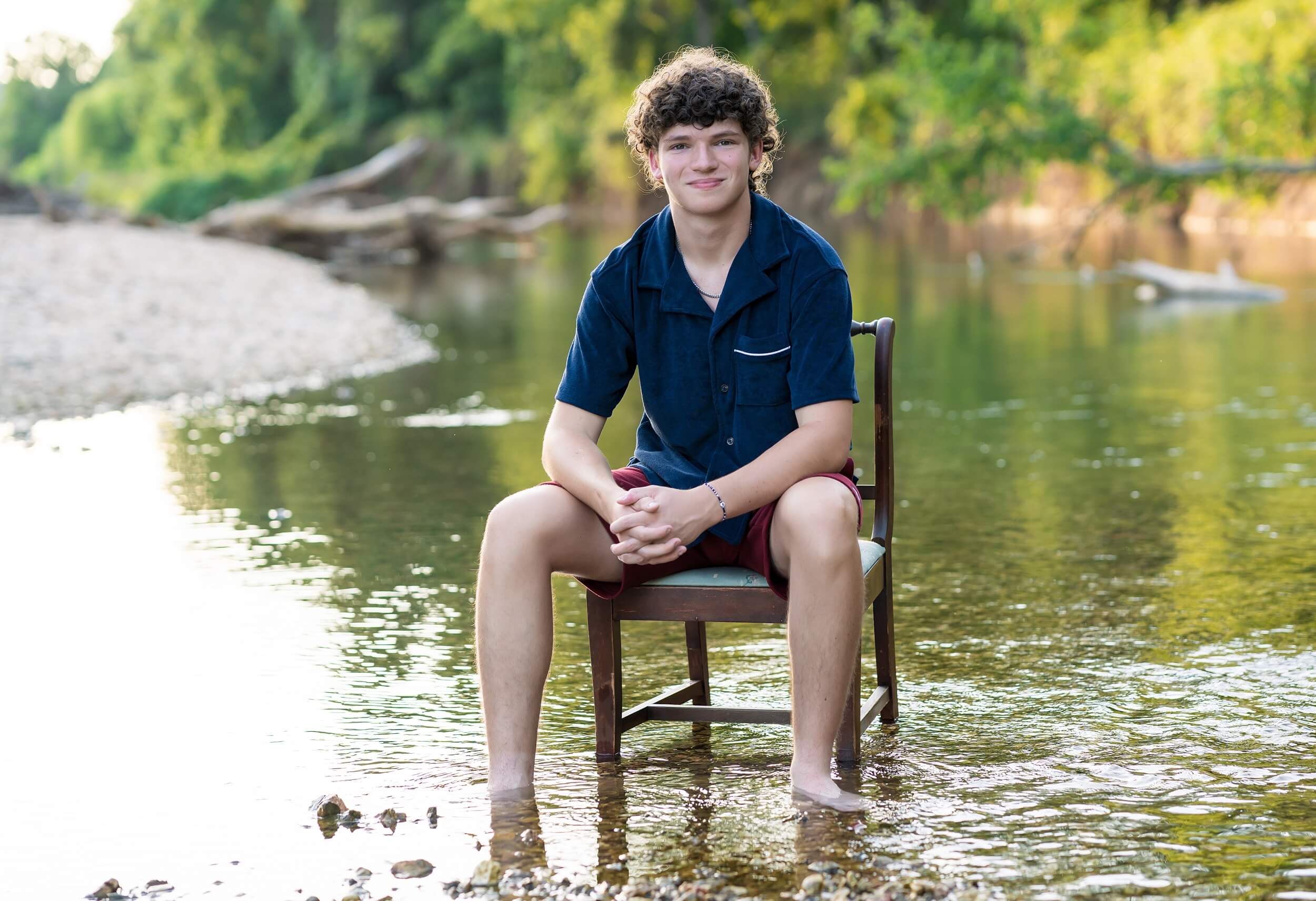 Example Shoot Title Tag A smiling high school senior sits in a wooden chair in a flowing river in a blue shirt leaning on his thighs