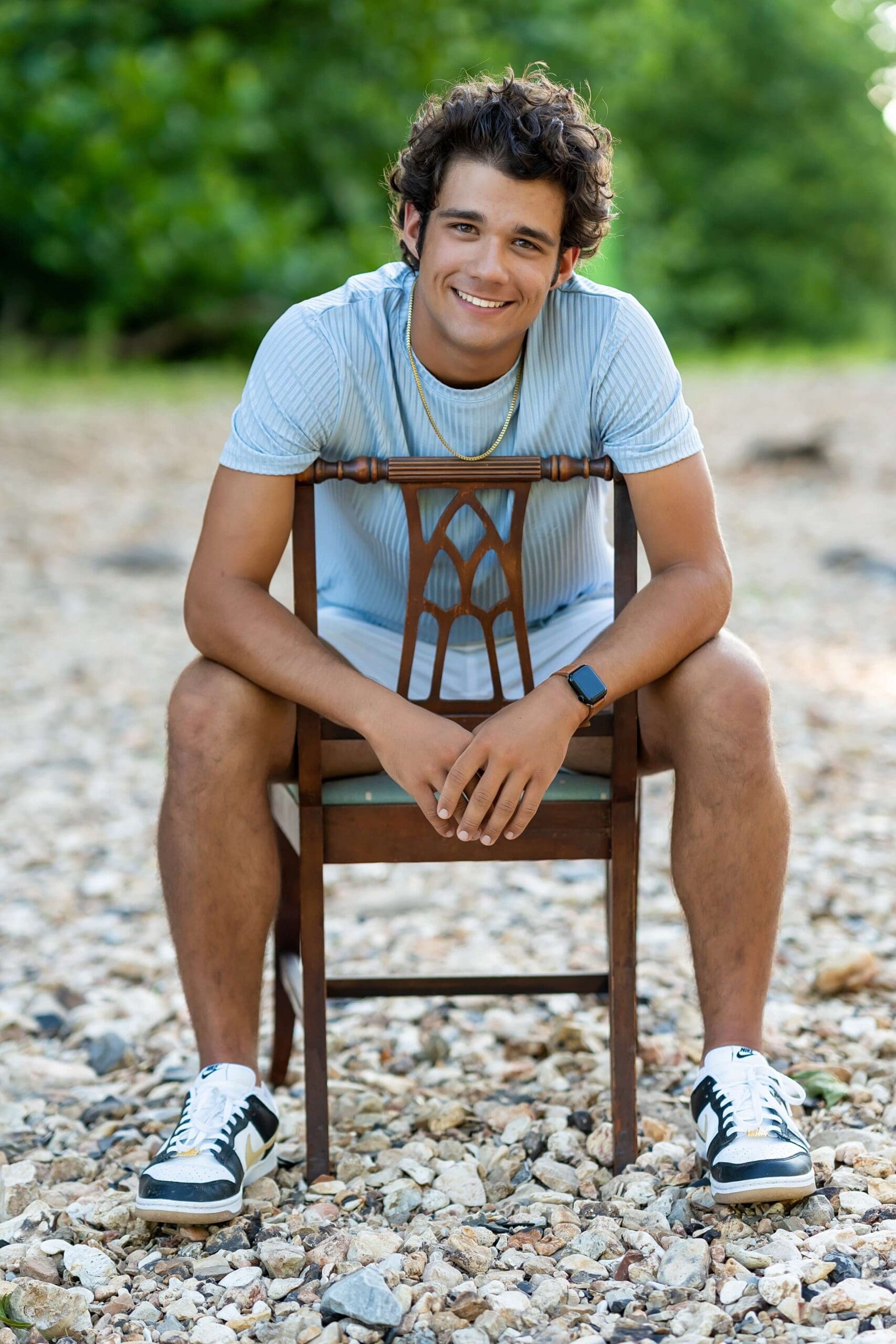 Example Shoot Title Tag A smiling high school senior sits backwards on a wooden chair on a rocky riverbed in a blue shirt