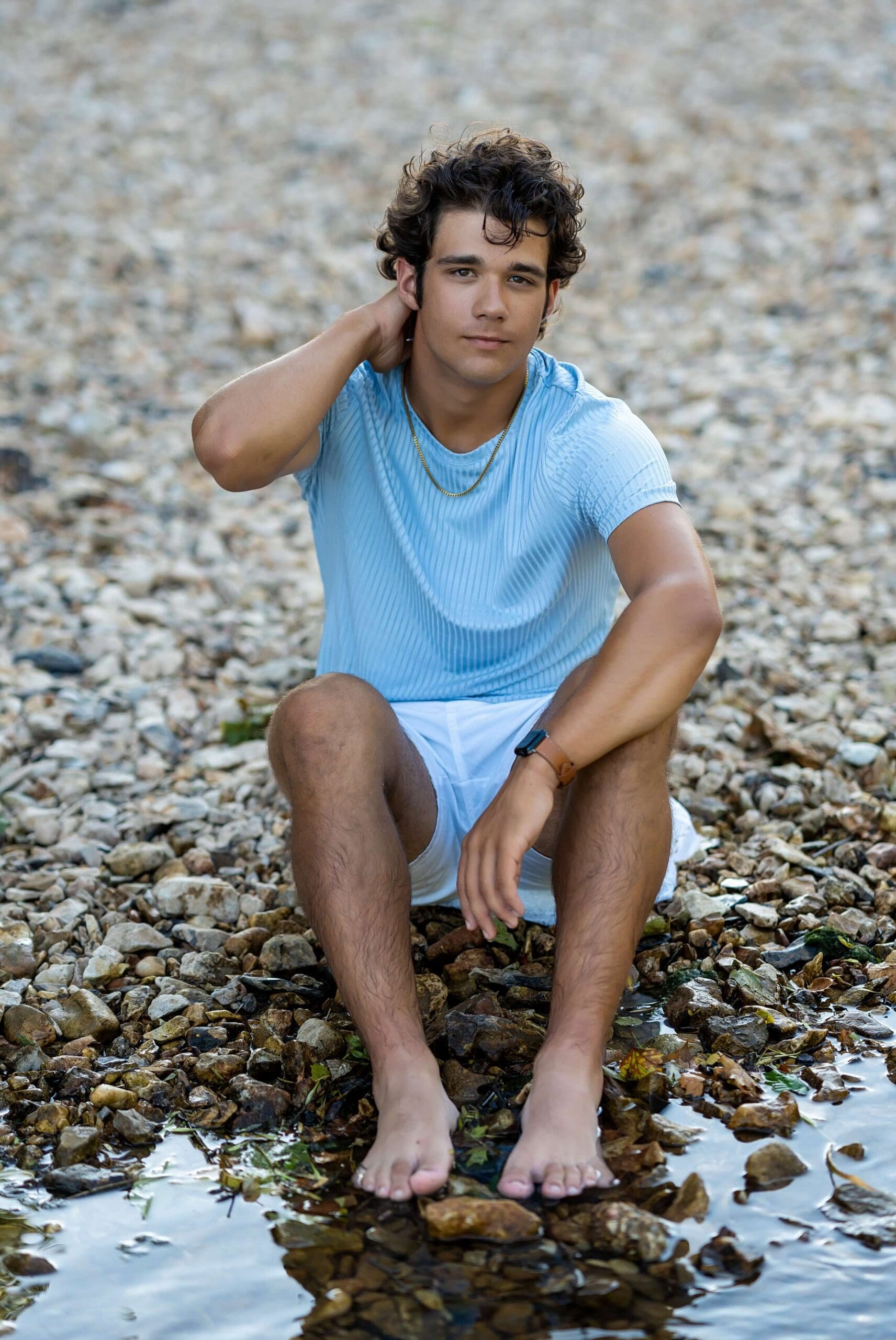 Example Shoot Title Tag A teenager in a blue shirt sits on the edge of a river pulling his hair back captured by an Arkansas senior photographer