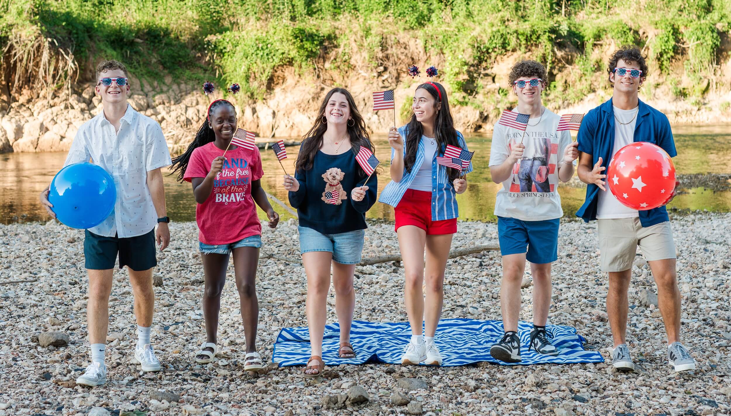 Example Shoot Title Tag Happy high school seniors walk on a rocky riverbed with big smiles, american flags, red white and blue balls and sunglasses