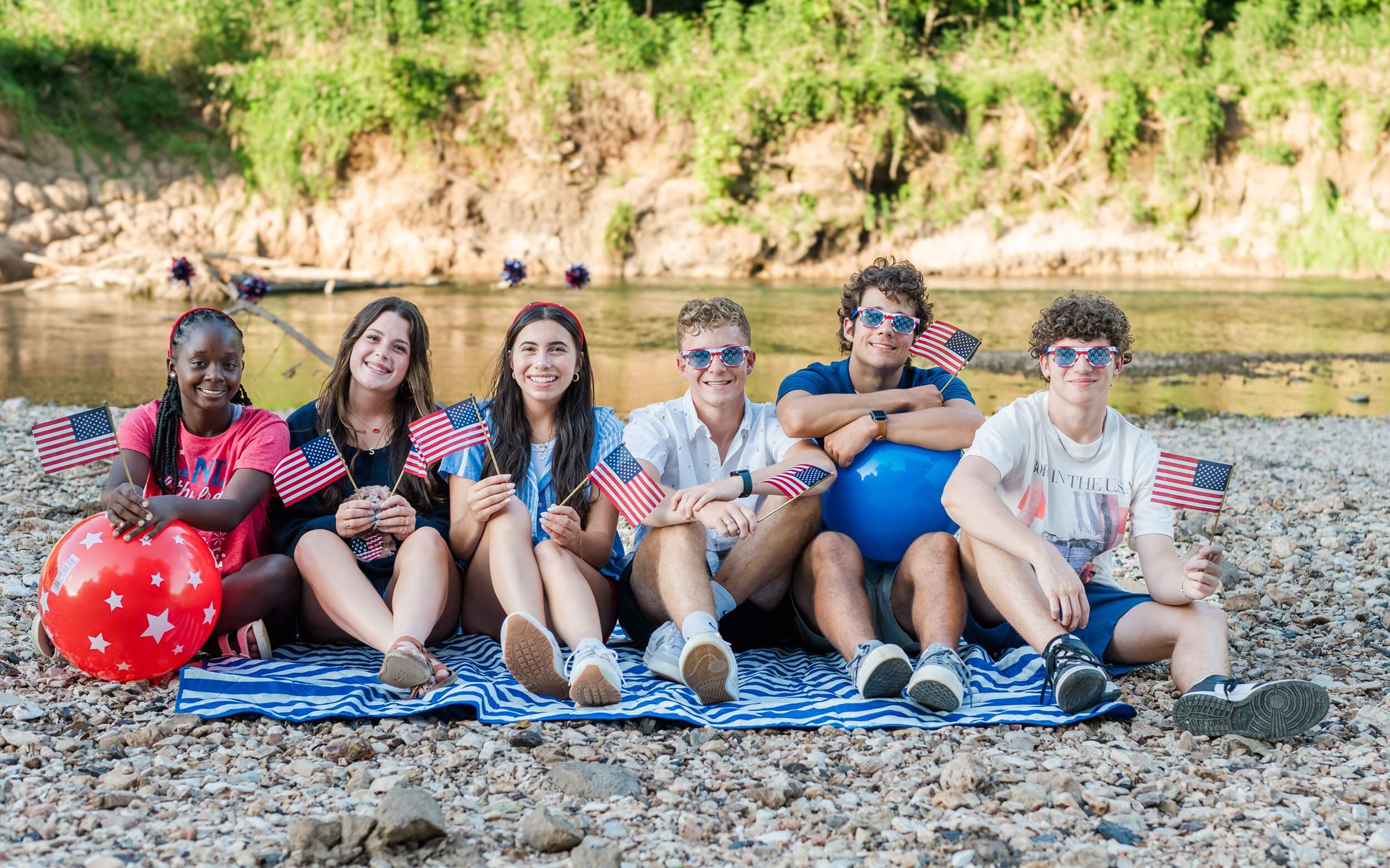 Example Shoot Title Tag A happy group of high school seniors sit on a towel on a riverbed sporting american flag sunglasses and flags