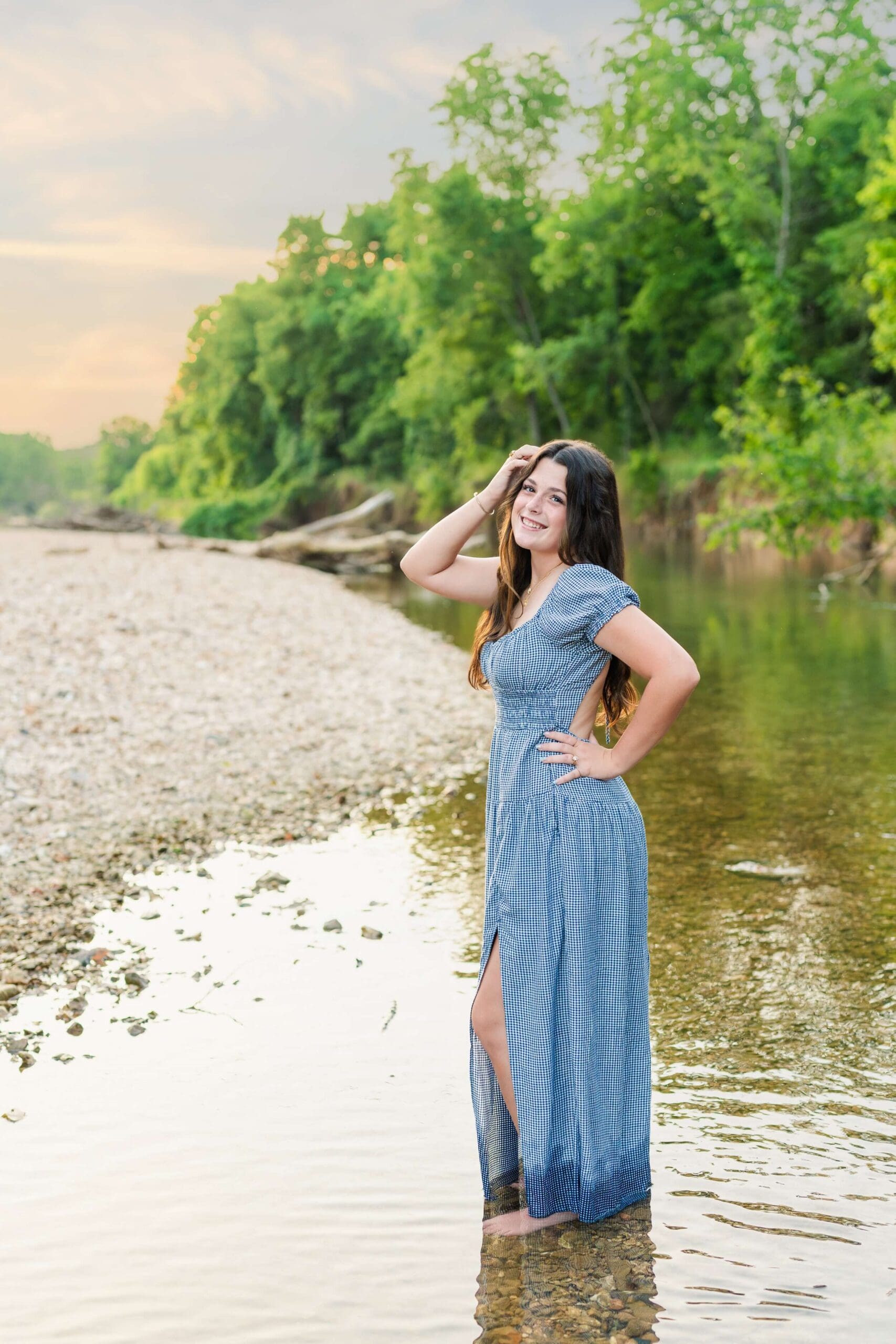 Example Shoot Title Tag A high school senior stands in a shallow river in a blue dress with a hand in her hair at sunset as guided by her Arkansas senior photographer