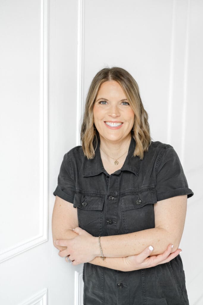 An Arkansas photographer standing against a white paneled wall with arms crossed, wearing a dark button‑up dress, photographed in a bright, minimal studio‑style setting.