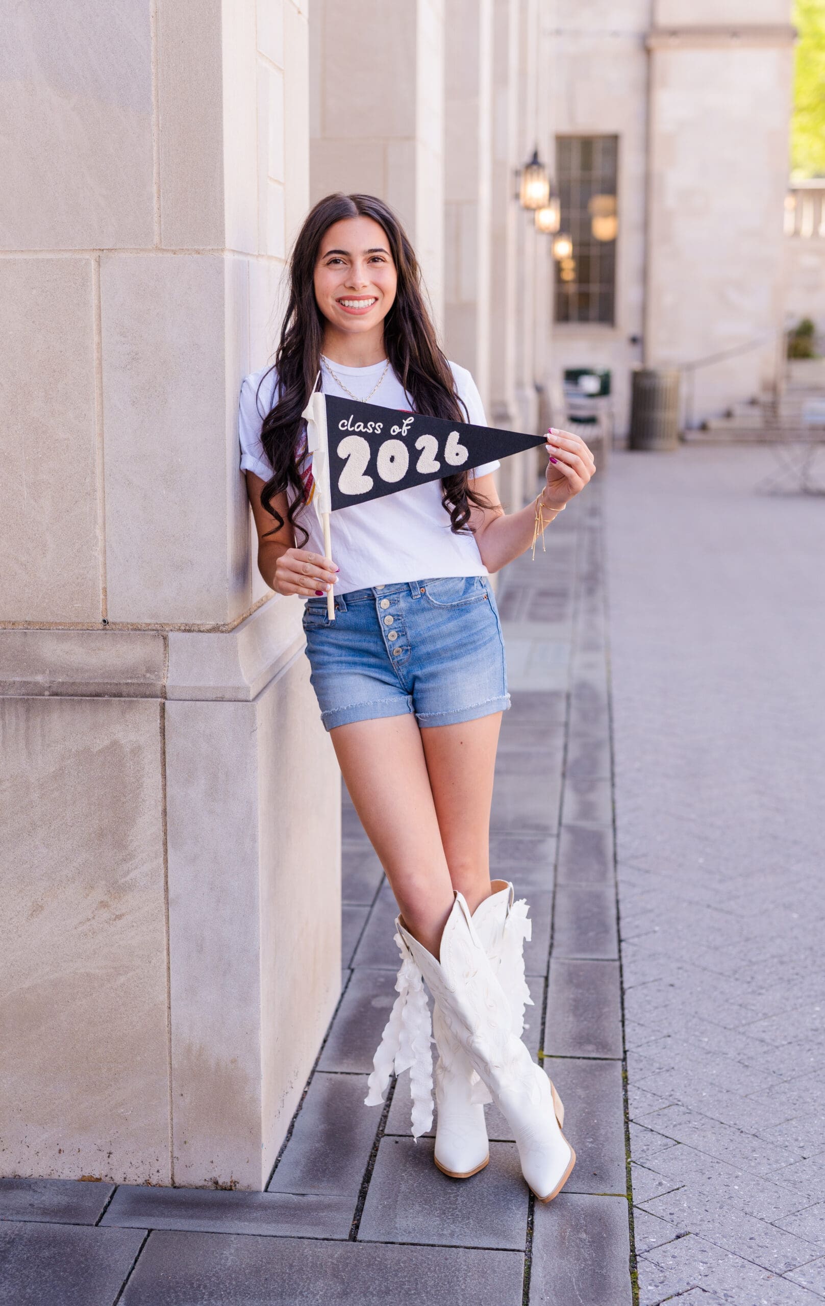 A high school senior holding a “Class of 2026” pennant while standing against a light stone building, photographed by an Arkansas Senior Photographer in a bright outdoor setting.